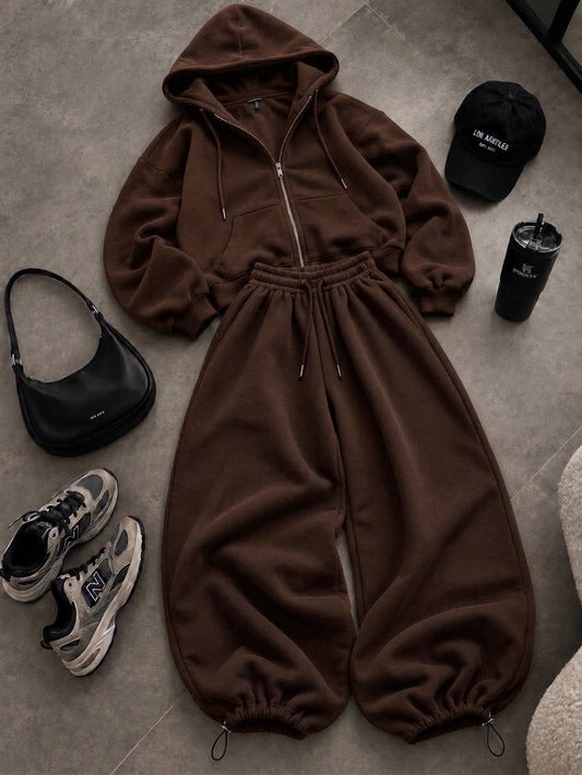 "06 oversized two-piece tracksuit featuring a zip-up hoodie with adjustable drawstrings and matching wide-leg sweatpants, styled on a grey floor alongside black accessories and sneakers."